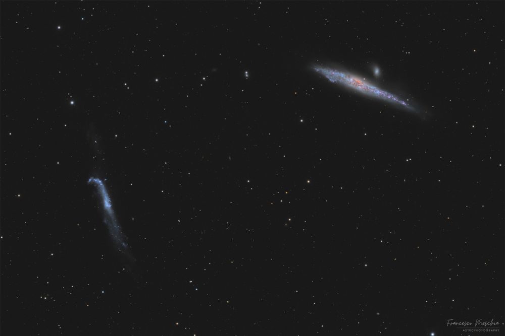 Photograph of two fuzzy galaxies. The one near the middle left resembles a hockey stick, and the one in the upper right resembles a stretched-out whale. The background is full of numerous stars and distant galaxies.