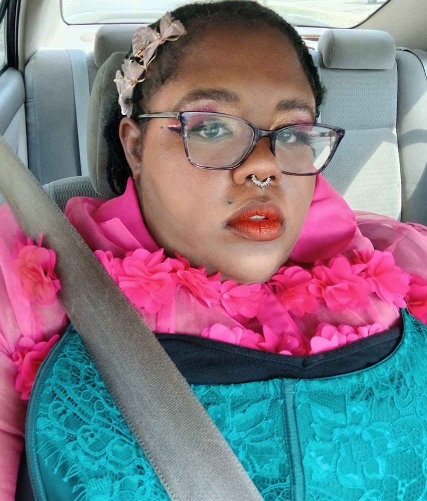Me with my glasses wearing a black dress with a pink flowery top underneath and a green lacy corset on top, with pink hair clips and my makeup done in a pink and red theme.