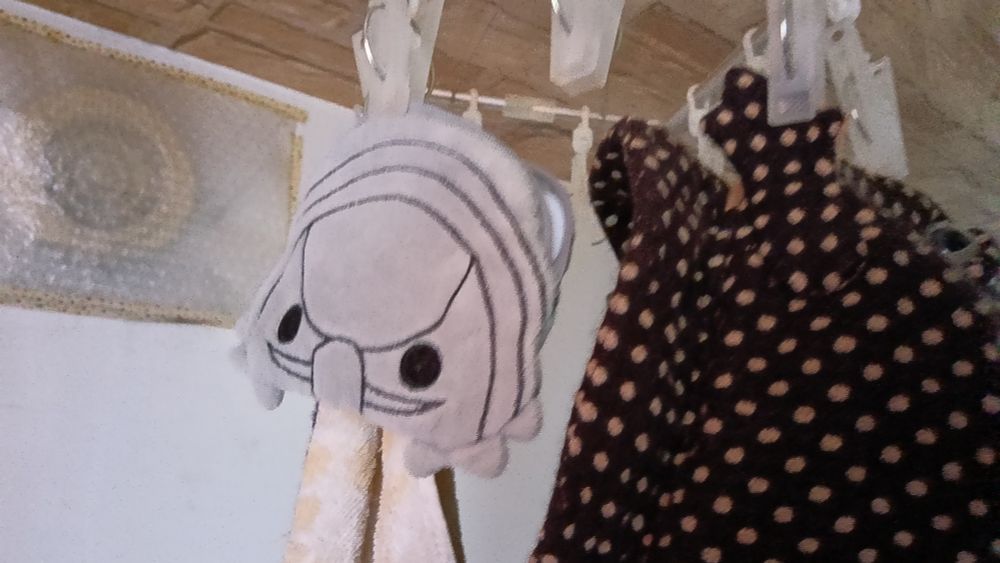 ピンチハンガーに、だいおうぐそくむしのぬいぐるみポーチを干している図です。全体が薄いグレーの生地で、背中部分にギャックがあり、開いた状態でピンチしています。右側にはふくらがえるを模したキャスケットの一部（焦げ茶にベージュの水玉の生地）が写っています。帽子の前面に顔と手がついており、両手をピンチして干しています。