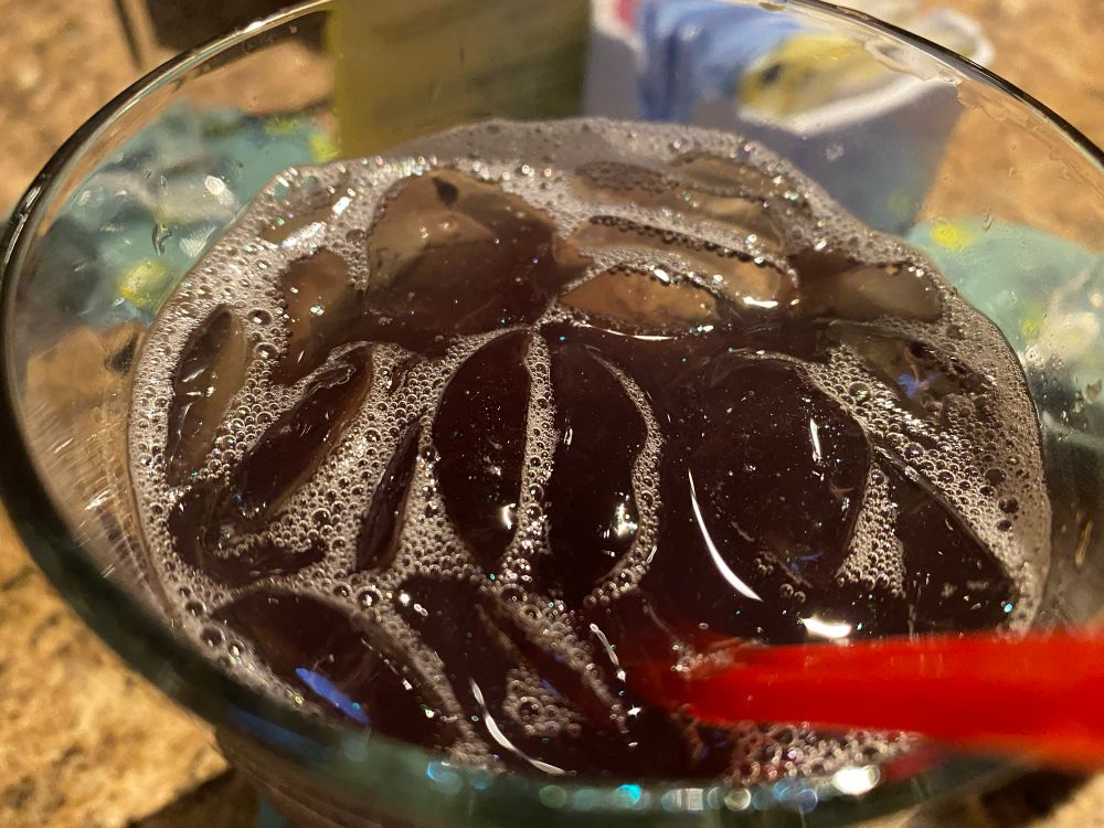 Close up on a dark colored cocktail with ice in it with edible glitter barely visible on top