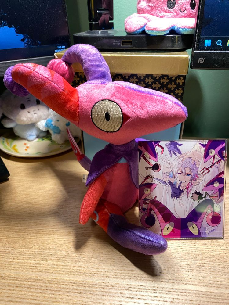 A photo of 2 pieces of Gavis Bettel's 2025 birthday merch sitting on top of a light colored wooden desk. On the left is a small plush of his mascot the Betelion, which is like a little pink and purple goblin with a very pointy head and horns. On the right is a rectangular acrylic coaster featuring art of Bettel in the center surrounded by Bettelion threatening him with various weapons