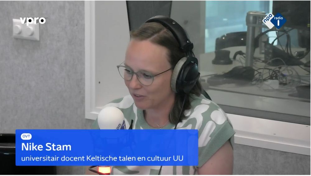 Nike Stam in front of a microphone with headphones on in the radio studio with a banner stating her name and function (universitair docent Keltische talen en cultuur UU)
