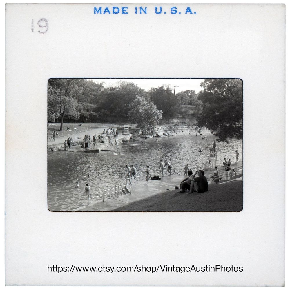 Black and white photograph of Barton Springs Pool in the 1950s, capturing a nostalgic summertime scene in Austin, Texas. The spring-fed pool is filled with swimmers of all ages, lounging on the grassy banks and wading in the cool water. Vintage swimsuits, modest by modern standards, reflect the era's fashion, while men and women relax on towels or perch on the stone edges of the pool. In the background, the natural limestone and tree-lined landscape of Zilker Park frames the scene, showcasing a time when Austin was still a sleepy town with fewer crowds. The photo evokes a sense of timeless community, summer joy, and the enduring appeal of Barton Springs as a local treasure.