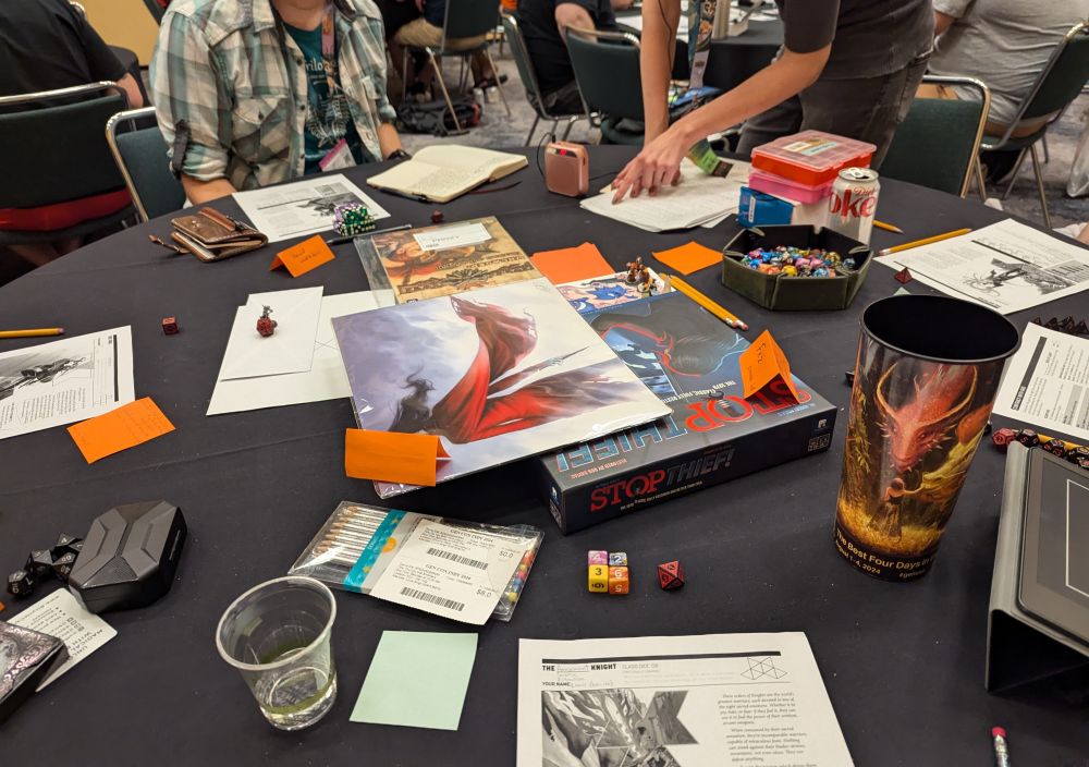 A table containing dice, character sheets (for the DIE RPG), and drinks. Taken at Gencon 2024.