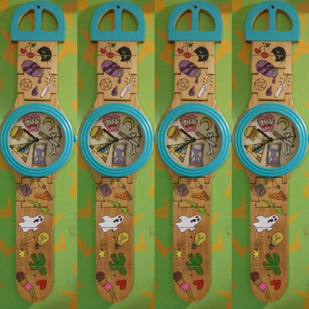 An oversize wooden wrist watch with a full size clock. Wood burned and painted doodles including ghost, cactus, ice-cream cone, lightbulb, pizza.
