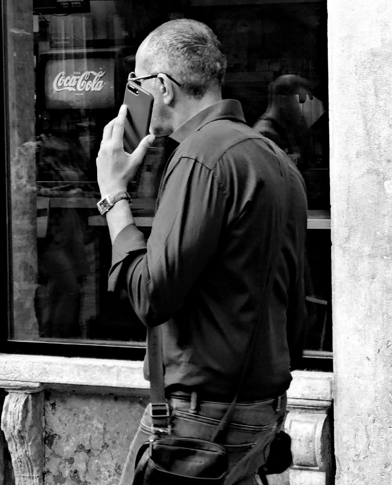 Black-and-white photo of a man talking on a cell phone. He is walking in front of a window of a small bar or restaurant with a Coca-Cola machine visible inside. There are numerous reflections in the window, including one of the man himself.