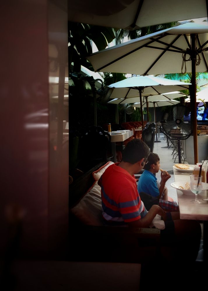Photo of a man and his son in an upscale outdoor restaurant. The father has what looks like a fancy cocktail and an elaborate entree; the boy is eating pizza.