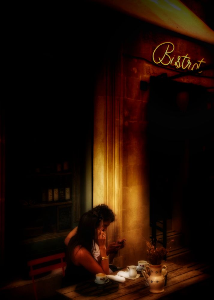 Photo of a couple having coffee at a table outside a bistro. The image mostly dark, with a warm golden light illuminating the couple. At first glance, it looks like the couple may be engaged in intimate conversation, but a closer look reveals that they are both on their cell phones.