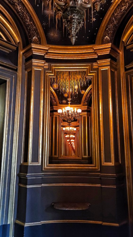 Photo of a chandelier in a round room of gilded mirrors, with the image reflected over and over in a receding line. The walls of the room are painted very dark blue with gold moldings, and the ceiling is painted with a gold sky and stars motif.