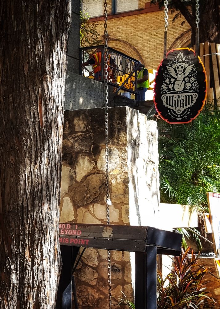 The side entrance of a rustic bar called The Worm, closed off for renovations. Two construction workers in bright vests are working on the balcony.