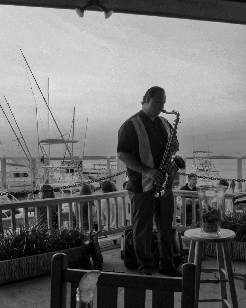 Black-and-white photo of a man playing a saxophone at an outdoor bar overlooking a marina.