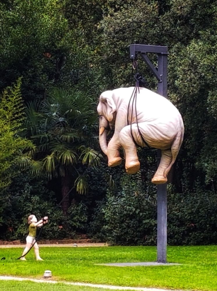 A lifesized sculpture of a child hoisting an elephant by a rope to the top of a post.