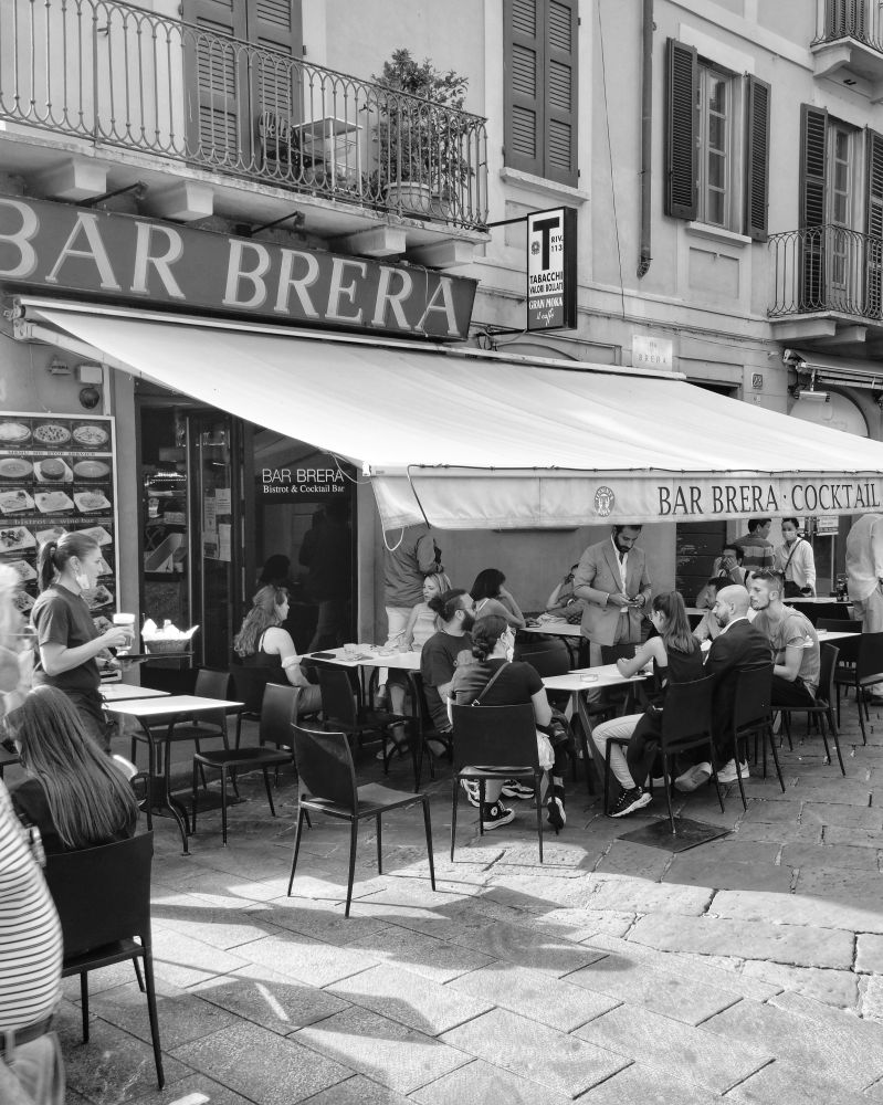 Black-and-white photo of a sidewalk bar with outdoor seating. There are numerous customers at the tables and several servers bringing food and drinks. 