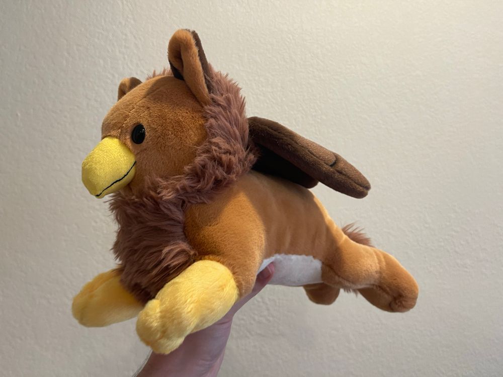 A brown, fuzzy Gryphon Plushie from Corviforms on BlueSky. It is facing to the left of the picture.