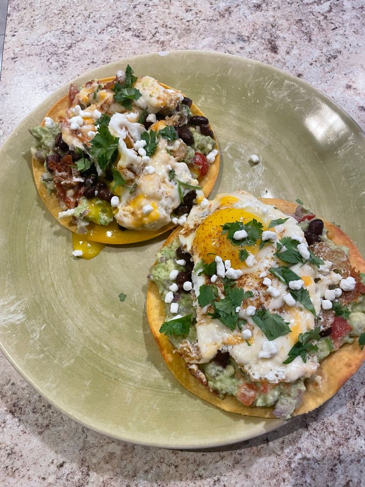 Two tostadas topped with homemade guac, black beans, a fried egg, cilantro, and goat cheese crumbles 