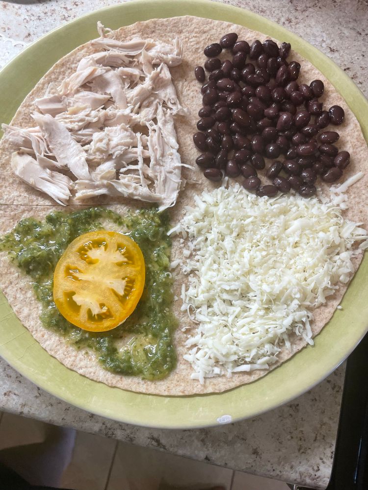 A tortilla on a plate with ingredients laid out in quarters. Top Left, shredded chicken; top right, black beans; bottom right, cotija cheese; and bottom left, salsa verde and tomato. 