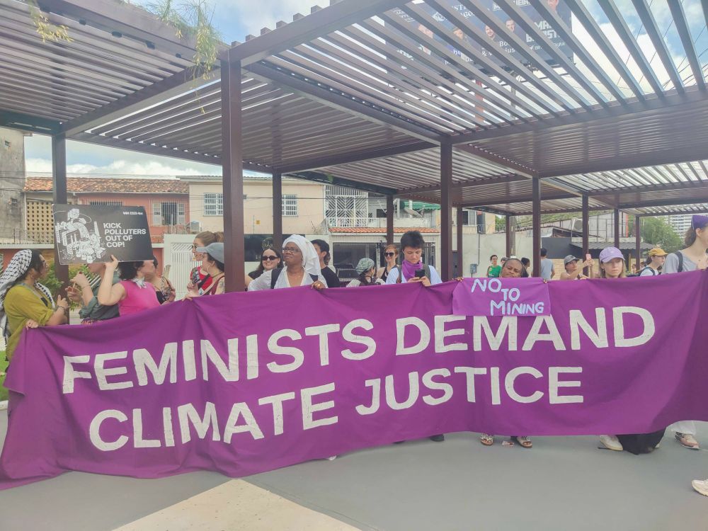 A diverse group of women stand together outdoors in front of a large purple banner reading “Feminists demand climate justice” and a smaller one "No to mining". 