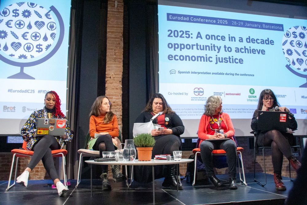 An all female panel on a stage. On the screen behind the panel is the tagline "2025: A once in a decade opportunity to achieve economic justice"