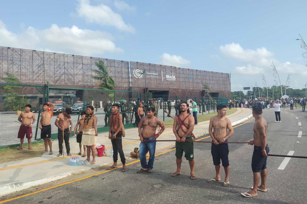 A group of Indigenous Peoples stands in a line on a street outside a fenced conference venue marked with United Nations and COP30 Brasil Amazonia signage. Behind the fence, security personnel are visible, and crowds gather further down the road.