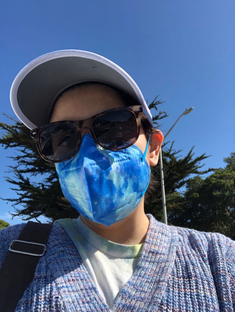 selfie of Meheen wearing sunglasses and a kn95 blue galaxy print mask and a purple baseball cap. also wearing a pastel blue sweater. outside there are some cypress trees in background.