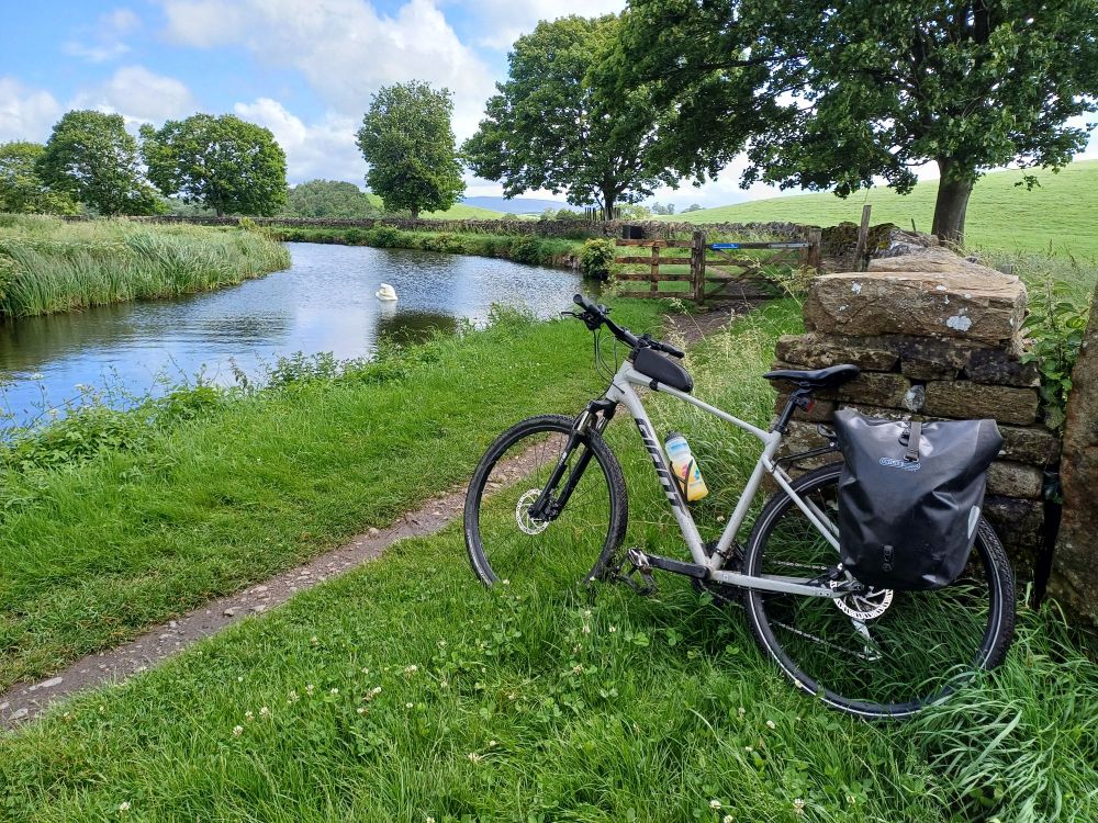 Photo of a cycle with a pannier leaning against va stone wall. A canal curves in from the left and away again. Trees, low hills and blue skies in the background.
