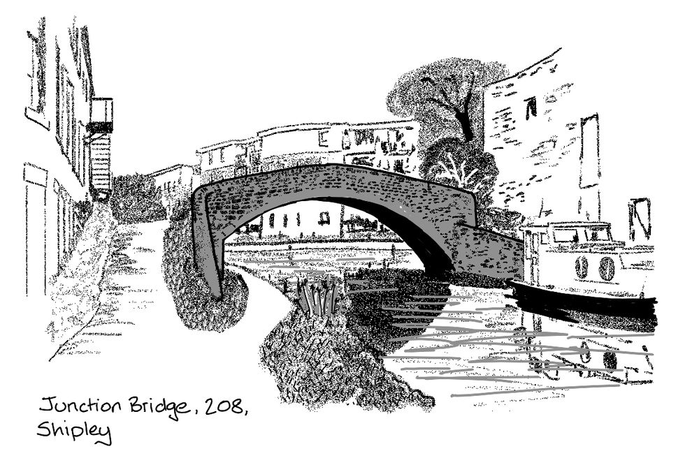 A stone bridge straddles a canal. To the left is the all of a factory, and to the right a boat and a brick wall. behind the bridge are some low rise blocks of flats.