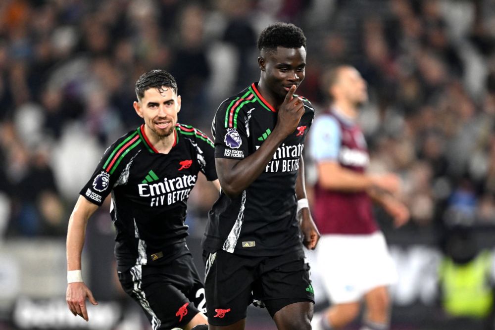 Saka celebrating after scoring his goal, putting his finger to his lips in a shushing gesture. Jorginho is running up to celebrate from behind him