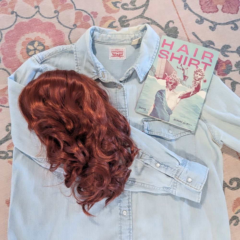 A light blue button down shirt rests on a cream rug with a floral pattern. There is a red curly wig on top of the shirt, as well as a copy of the poetry collection "Hair Shirt" by Adrian Sobol.