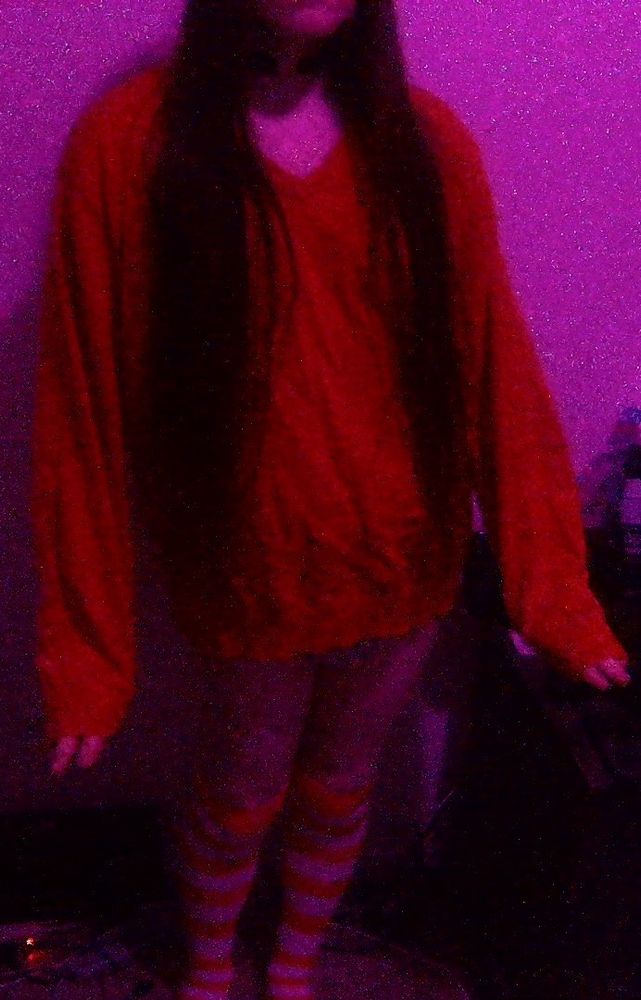 An absolutely adorable person with long brown hair, wearing a choker with a heart emblem, orange sweater, and orange and white stripped thigh highs. Off screen, they are wearing a pair of glasses. They're striking a cute pose.