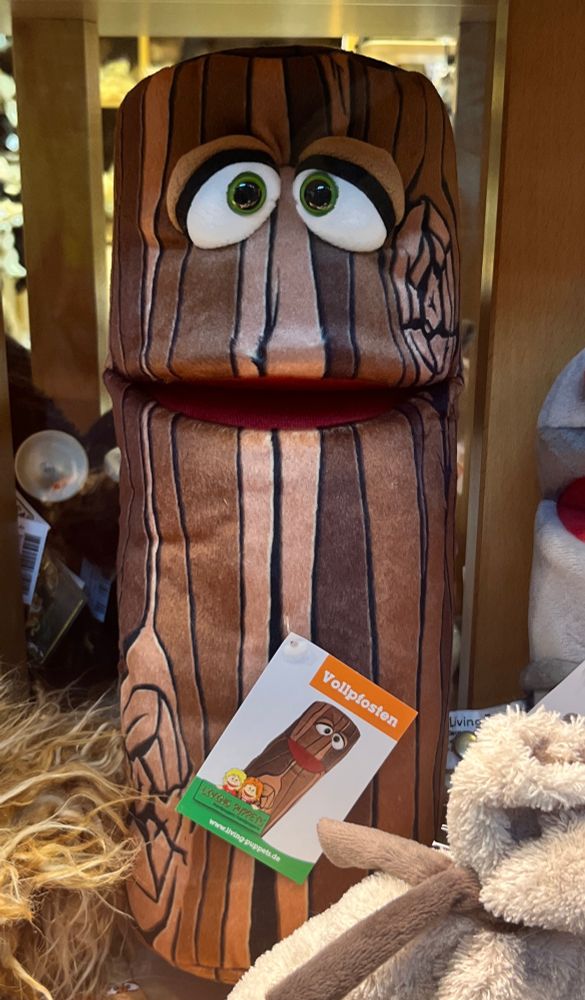 Kuscheltier, im Prinzip ein Stamm mit Augen und Mund. Dazu ein Pappschild, auf dem „Vollpfosten“ steht.