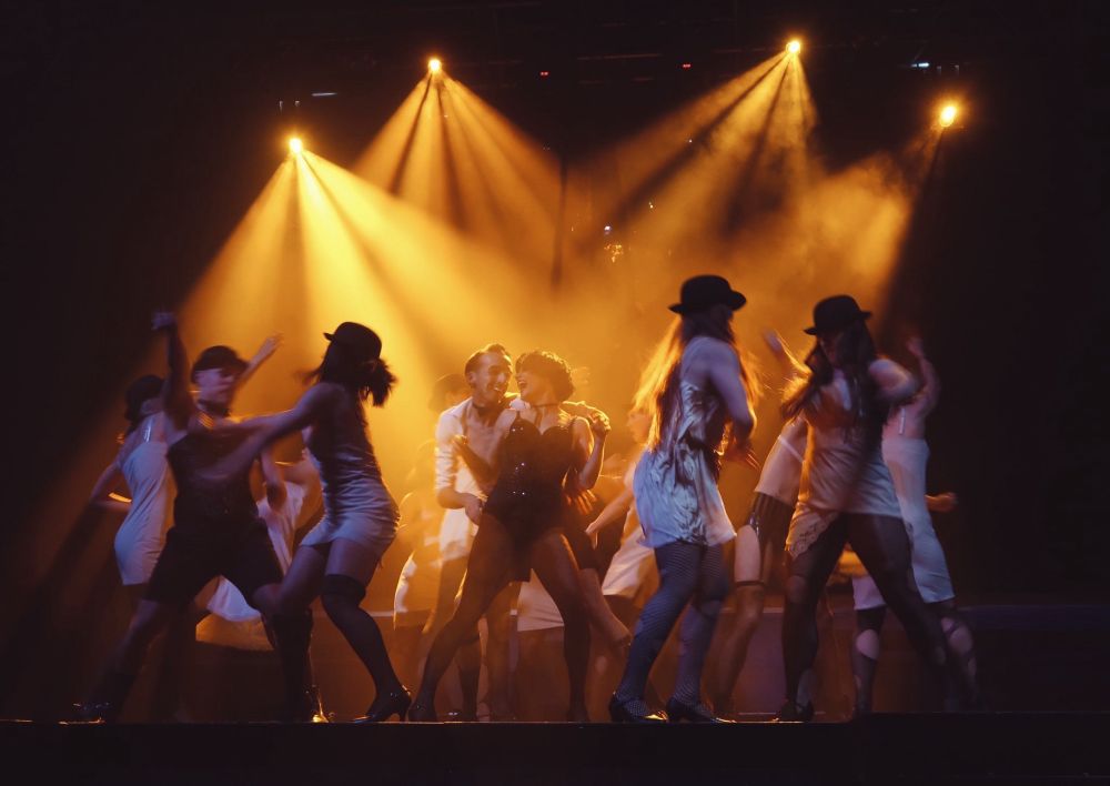 A group of dancers around a central couple, with movement. Yellow broken up lighting from behind 