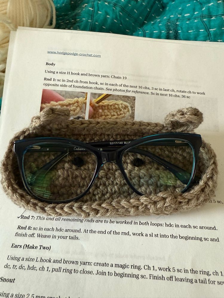 My blue-framed glasses in an oblong brown crocheted glasses holder. The bear has ears and beady little eyes but she's missing her snout and nose. It's sitting on top of a printed copy of the pattern which is sitting on a teal blanket with a ball of white yarn.