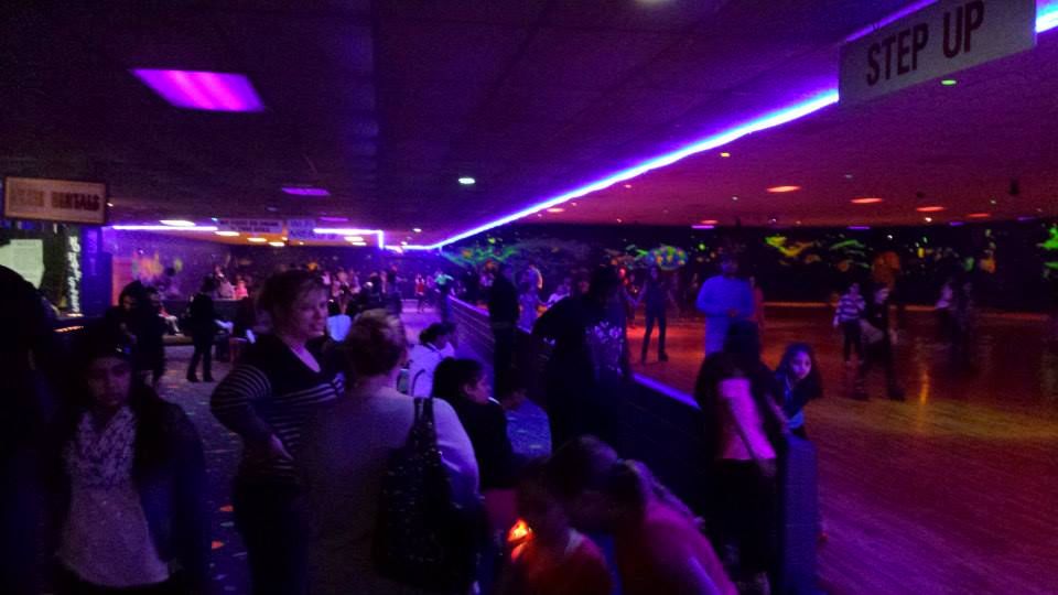 A crowd, bathed in purple neon light, is gathered at the edge of the skating floor at Roller World roller rink in Saugus, Massachusetts.