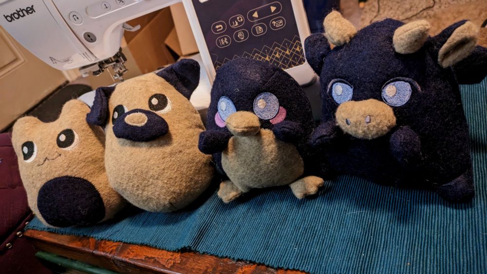 Photo of four small plushies in a line on a wooden table with a white sewing machine behind them. The plushes are made of tan and blue fabric with embroidered eyes. From left to right the plushes are a cat, dog, penguin, and dragon