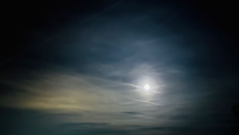 A half moon glows softly behind thin clouds in a dark night sky, surrounded by gentle shades of blue and gray, creating a peaceful and mysterious mood.