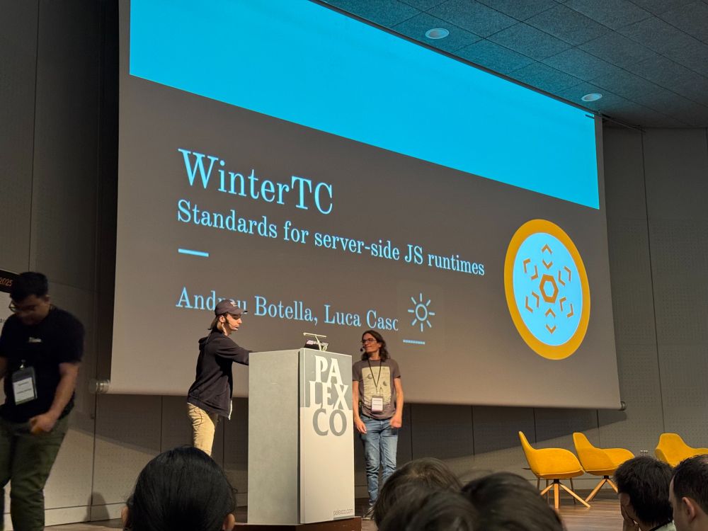 Two people standing at the podium in front of a large screen and auditorium getting ready to talk about WinterTC, Standards for server-side JS runtimes.