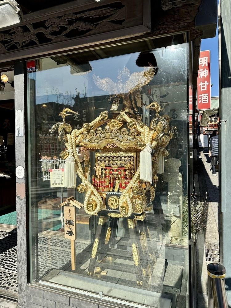 A gilded and ornate portable shrine is displayed in a glass-fronted case on a sunny street.