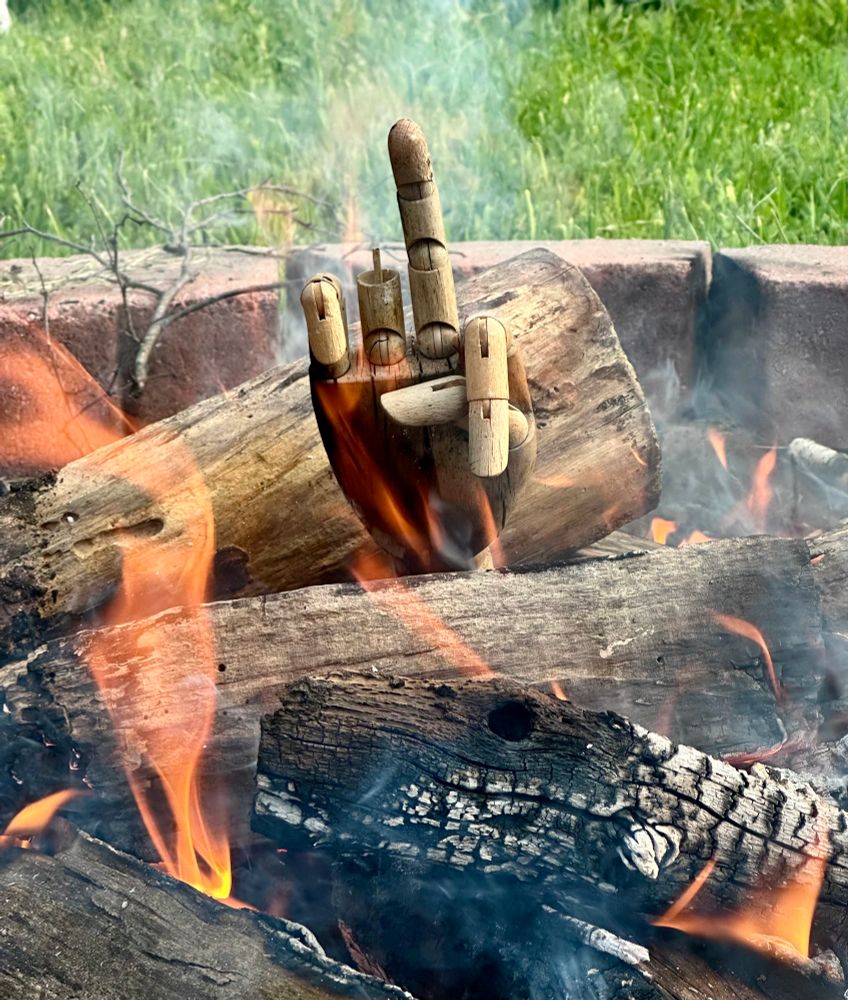 A bonfire in a brick-ringed fire pit with a broken wooden hand model sticking up out of the middle of the fire with its middle finger extended.
