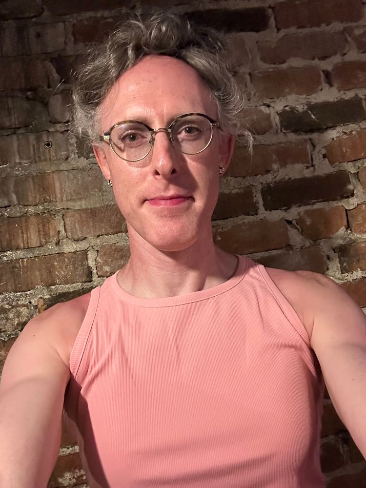 A selfie of Bee, a trans woman, seen from the chest up. She is wearing a soft pink high-neck tank dress, and her hair is up in a very messy bun. Behind her is an old brick wall.