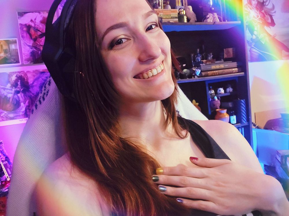 Strixxline smiles at the camera with a hand to their chest. Their nails are painted in rainbow colors.