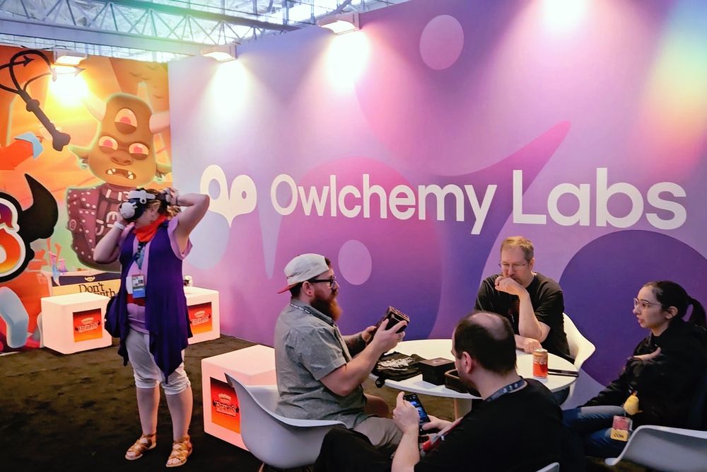 Folks sit at a table in front of a backdrop with the Owlchemy Labs logo and wordmark over a purple background. Nearby, one person is putting on a VR headset. On the other wall is another backdrop featuring art from their game Hexas.