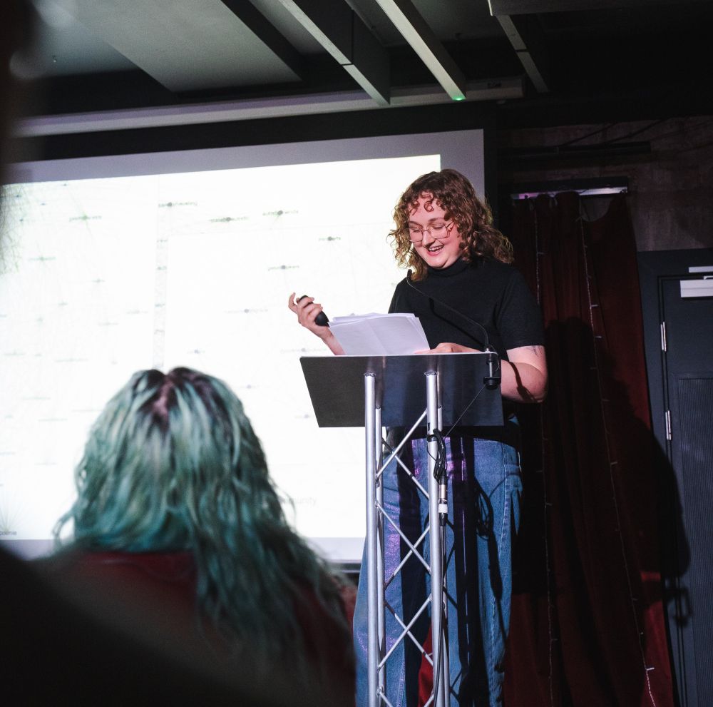 Meg (they/them) is standing at a lectern looking down at their notes and laughing. They're wearing a short-sleeved black turtleneck and iridescent-pink blue jeans. Behind them is a PowerPoint slide showing a close-up of some network graph data. A person with mid-length turquoise hair sits in front of the camera to the left of Meg, watching their presentation.