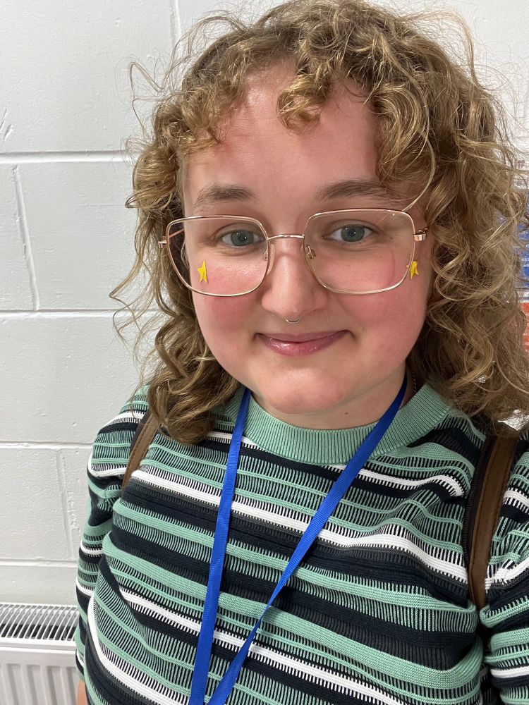 A selfie of Meg, a white person in their late twenties, wearing a teal black and white stripy top, square glasses and a gold star sticker on each cheekbone. Meg is smiling happily at the camera as they’ve just left an amazing panel discussion on neuroqueer performance at the Critical Neurodiversity Conference.