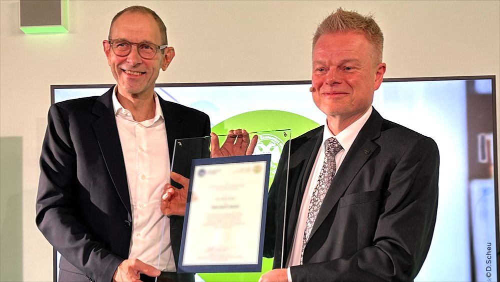 Profesor Ulrich Thonemann, Ph.D. and André Drost Head of the WIso Library and camous Servidce in suits holding up a certificate encased between two clear acrylic panels during an award ceremony. Thonemman, wearing glasses and a white shirt, smiles at the camera. André Drost on the right, wearing a suit and a patterned tie, also smiles. The certificate, held centrally is displayed in front of a screen with a green and white graphic.