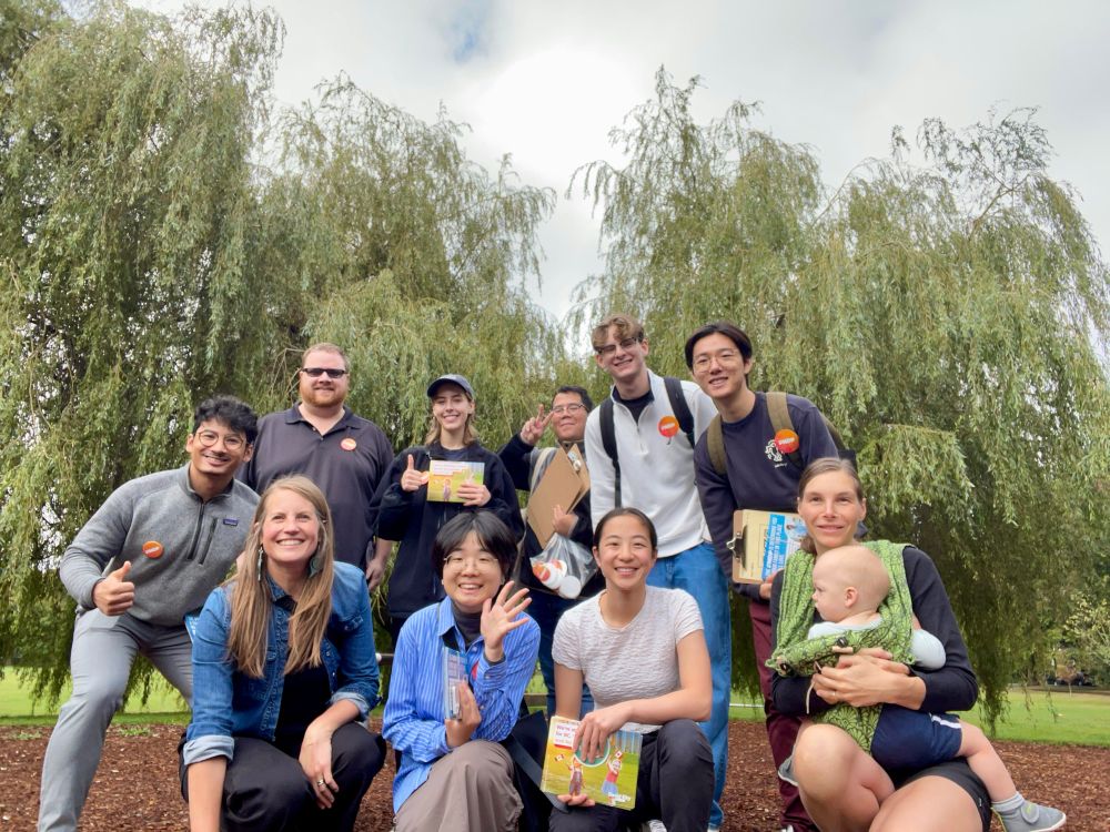 Photo of ten volunteers and a baby gathered together with flyers and clipboards before going out door knocking. They are in a park with Willow trees behind. 