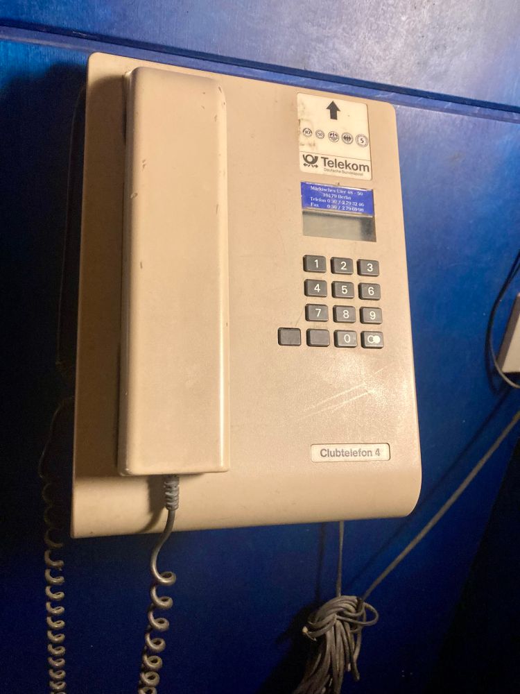 Vor nachtblauer Wand ein als Clubtelefon ausgewiesenes Fernsprechgerät der Telekom mit Tasten und einem Einwufschlitz für Münzgeld. 