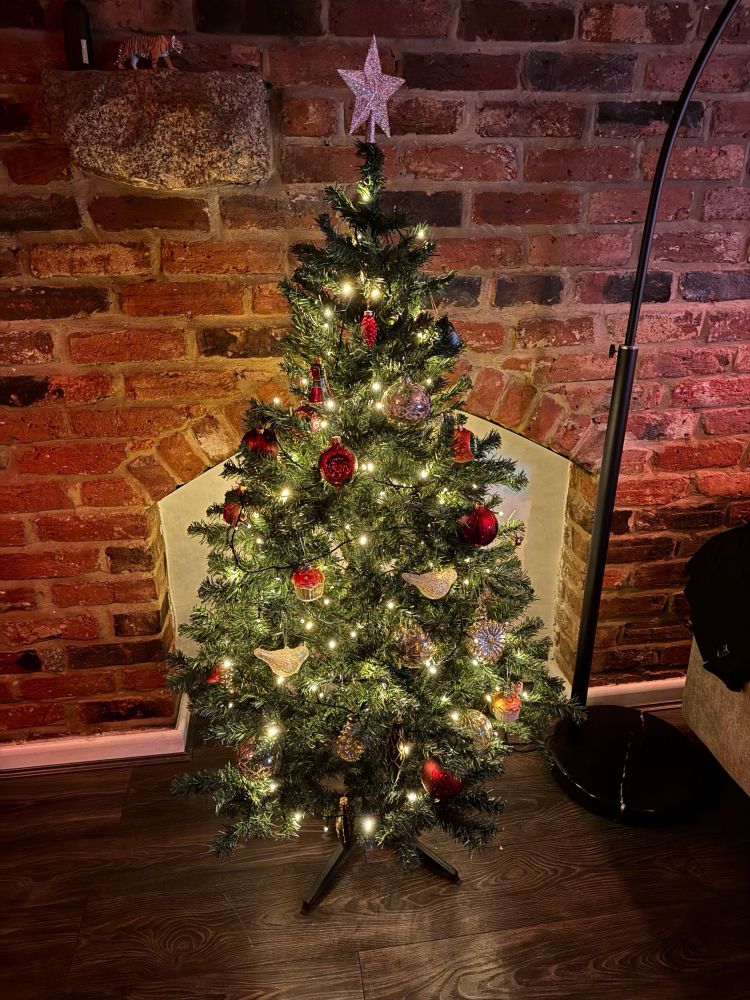 Artificial Christmas tree, decorated with white, red, and green decorations made of glass. In a living room with a brick wall and cozy warm white light.