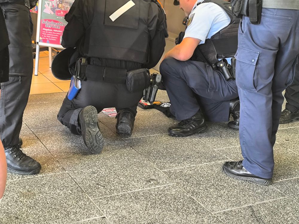 Man sieht verschiedene Gegenstände auf dem Boden liegen, darunter auch welche mit schwarzer Sonne und Reichsfarben. Davor knien Cops und schauen sich diese an