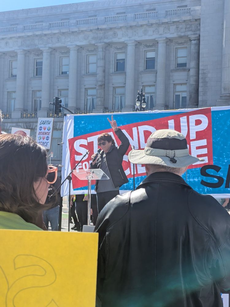 Carolyn Bertozzi stands at a podium with two fingers up in the air. Crowd in the forefront and background with signs supporting science.