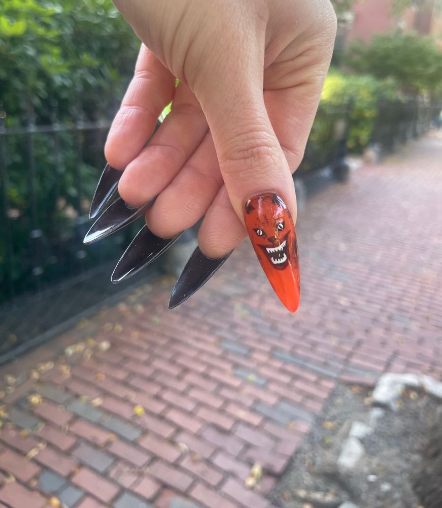 Darja shows off the hand painted art on her stiletto manicure – the thumb is a perfect rendition of the cat on the movie poster for House (1977) and the rest are dark brown. Greenery and Boston cobblestone streets are visible behind.